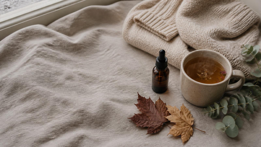 Amber glass serum bottle on a linen throw beside a mug of herbal tea, a cream knit jumper, autumn leaves and eucalyptus.