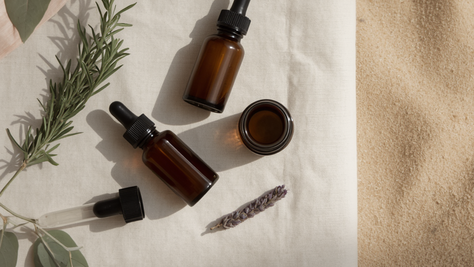 Amber glass dropper bottles and a small amber jar arranged on pale linen with sprigs of rosemary, eucalyptus and dried lavender, beside soft coastal sand in warm natural light.