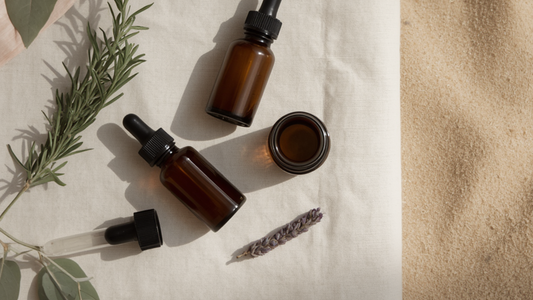 Amber glass dropper bottles and a small amber jar arranged on pale linen with sprigs of rosemary, eucalyptus and dried lavender, beside soft coastal sand in warm natural light.