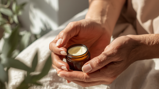 Mature woman's hands gently holding an open amber glass jar of natural cream, with soft sunlight and botanical shadows in the background